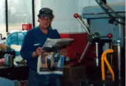 FL Decker wearing a hat and reading a newspaper in a machine shop